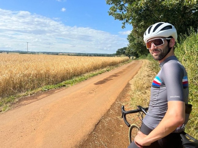 James Zuccollo leaning on his bike during a bike ride next to a field.
